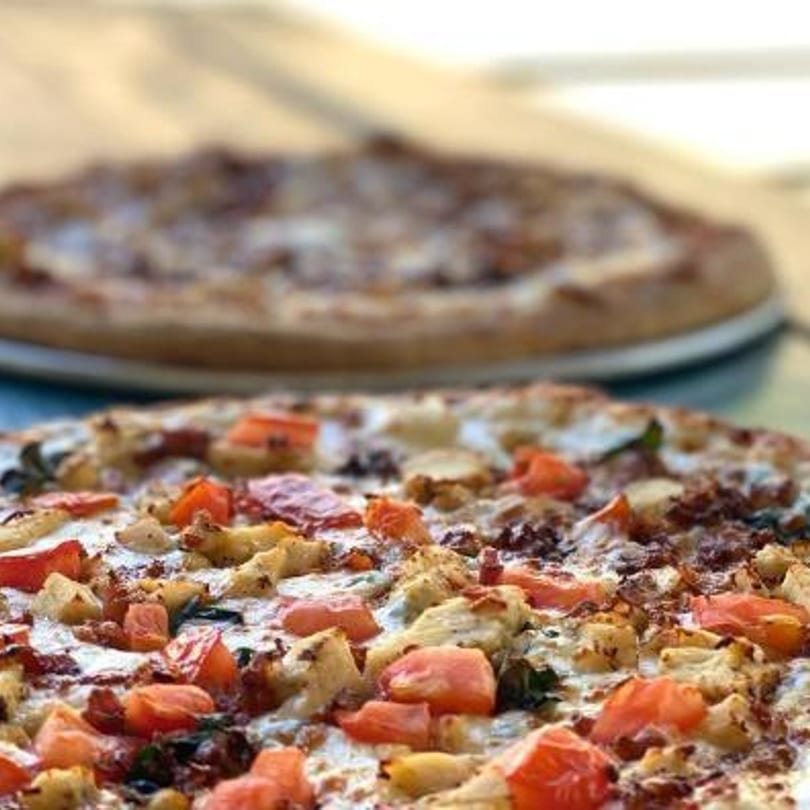 Two Gumby's pizzas, one in focus with chicken, tomatoes, and white sauce, another blurred in the background.