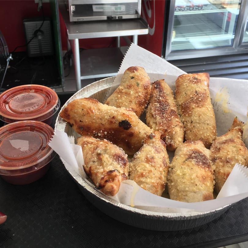 A foil container of golden-brown Gumby's Pizza pepperoni rolls with two cups of marinara sauce.